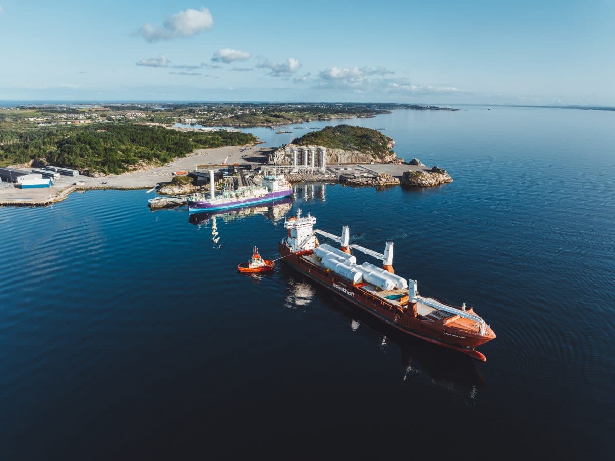 Flyfoto av et lasteskip og en slepebåt i nærheten av en havn med industribygninger og en kystlinje i bakgrunnen.