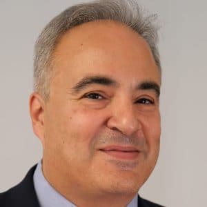 En middelaldrende mann med kort, grått hår og et lite smil, iført mørk dress og lyseblå skjorte, fotografert mot en enkel, lys bakgrunn.