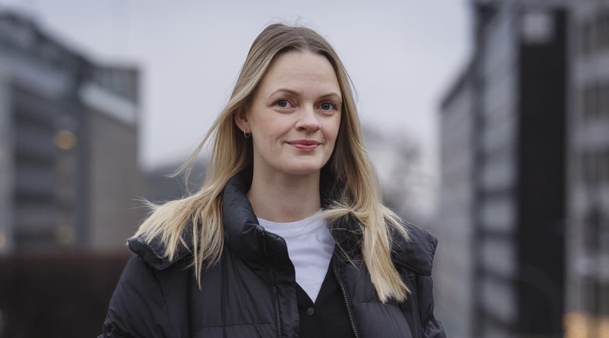 En kvinne med blondt hår i svart jakke og hvit skjorte står utendørs i et urbant område med bygninger i bakgrunnen.