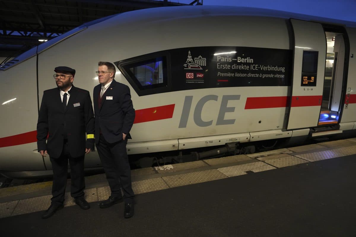 To konduktører står på perrongen ved siden av et hvitt ICE-høyhastighetstog, merket med et skilt for direkteruten Paris-Berlin, på en togstasjon i skumringen.