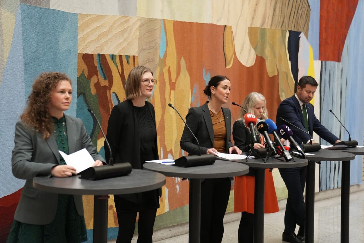 Fem personer står bak podier med mikrofoner og snakker på en pressekonferanse foran et fargerikt, abstrakt veggmaleri.