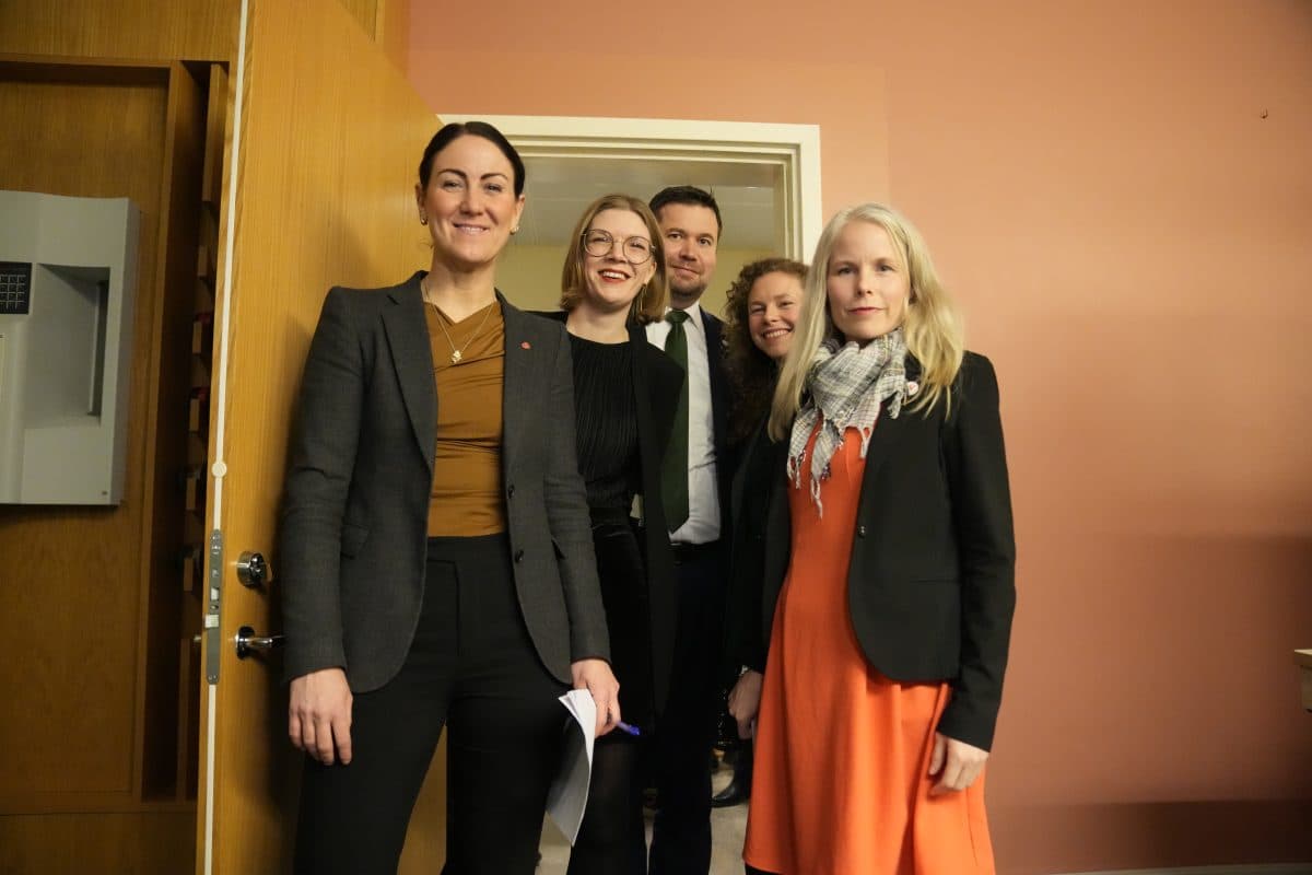 Fem voksne mennesker, tre kvinner foran og to bak, står smilende sammen i et rom med rosa vegger og en åpen tredør.