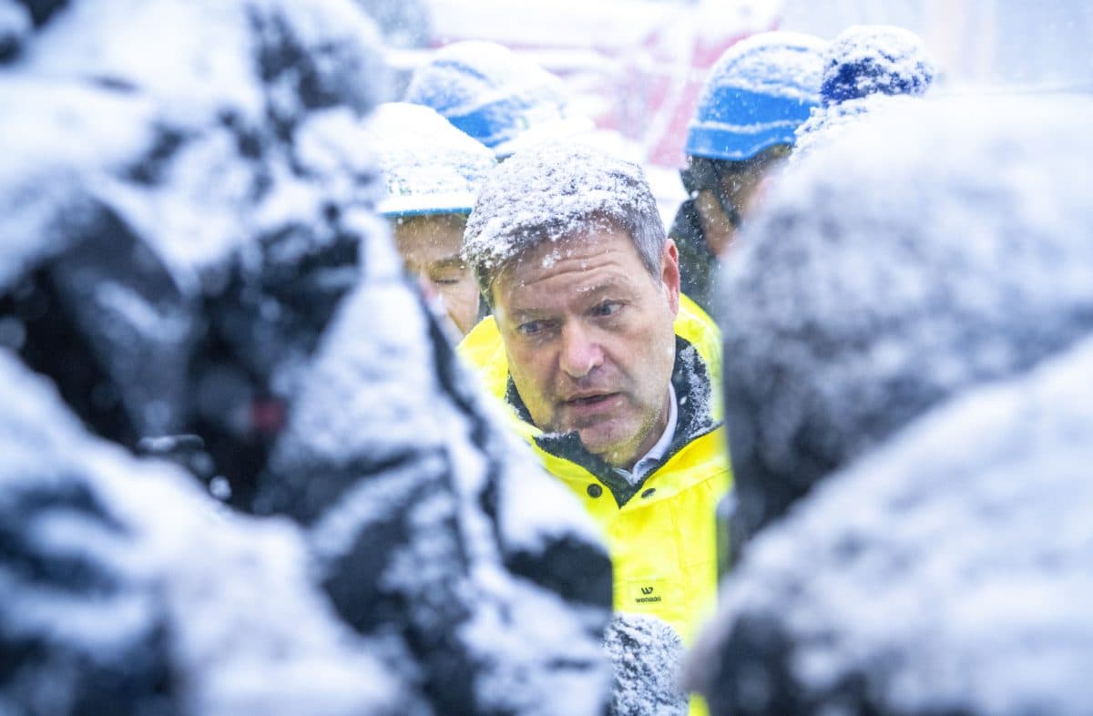En mann i gul jakke står utendørs blant hjelmkledde mennesker, omgitt av kraftig snøfall og snødekte flater.