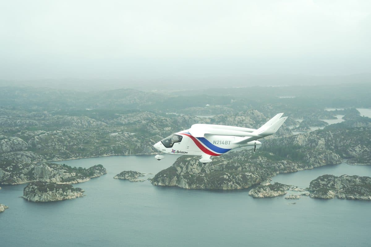 Et hvitt, elektrisk fly med røde og blå aksenter flyr over et tåkete landskap av steinete øyer og vann.