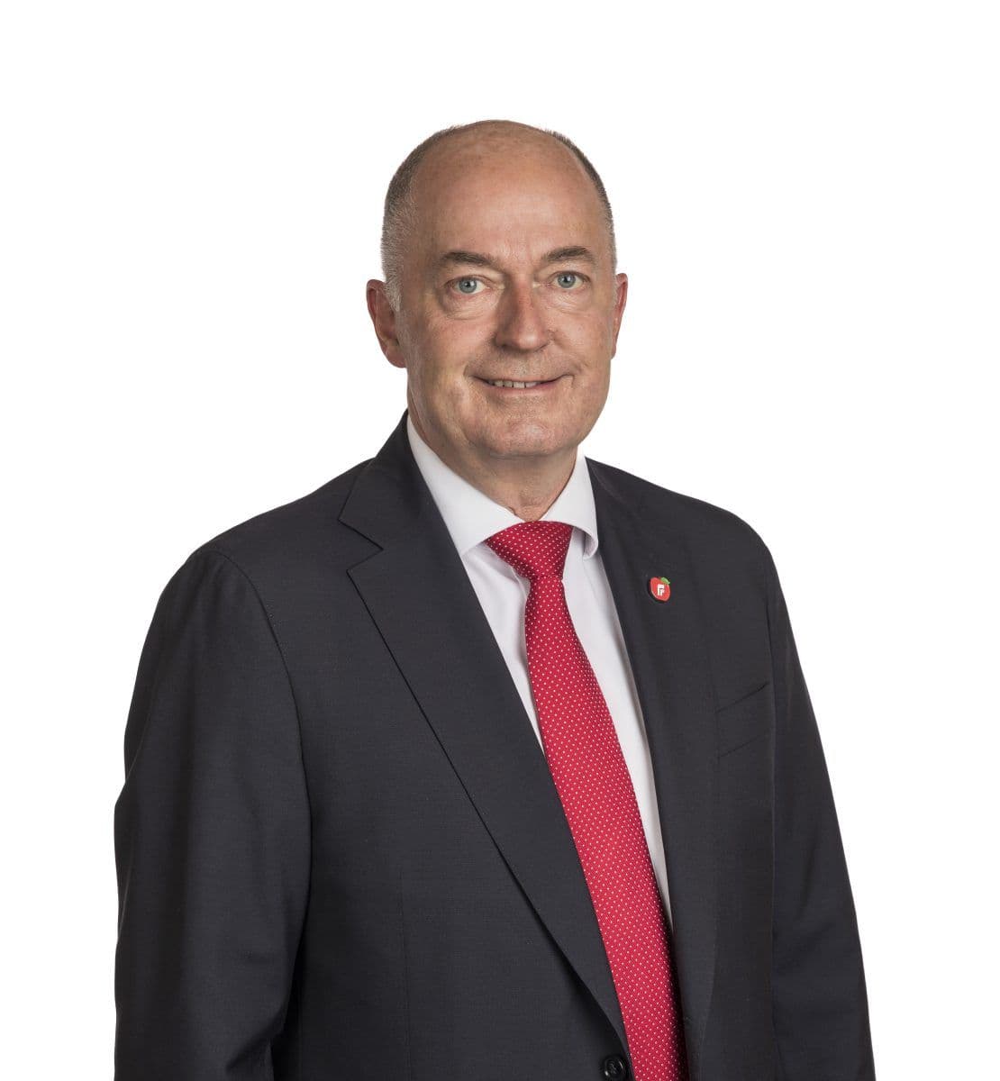 En middelaldrende mann i mørk dress, hvit skjorte og rødt slips står mot en vanlig hvit bakgrunn.