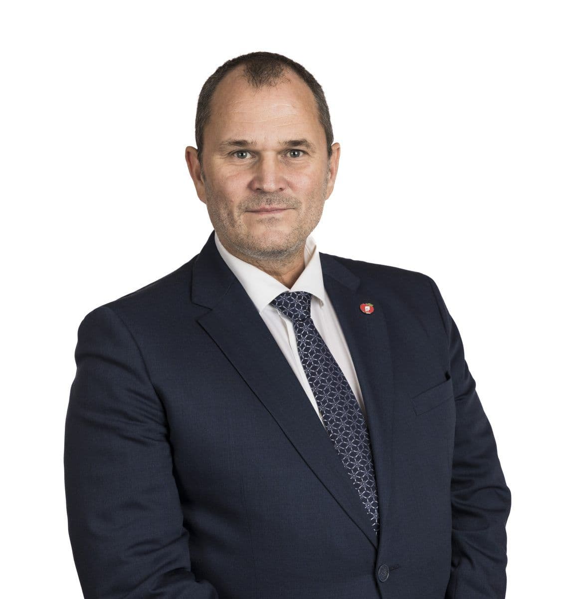 En mann i marineblå dress, hvit skjorte og mønstret slips poserer mot en ensfarget hvit bakgrunn. Han har kort hår og bærer en jakkeslagsnål.