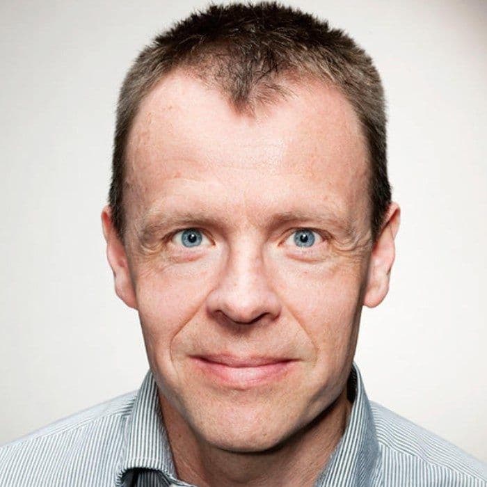 En person med kort hår iført en stripet skjorte, ser direkte på kameraet mot en vanlig bakgrunn.