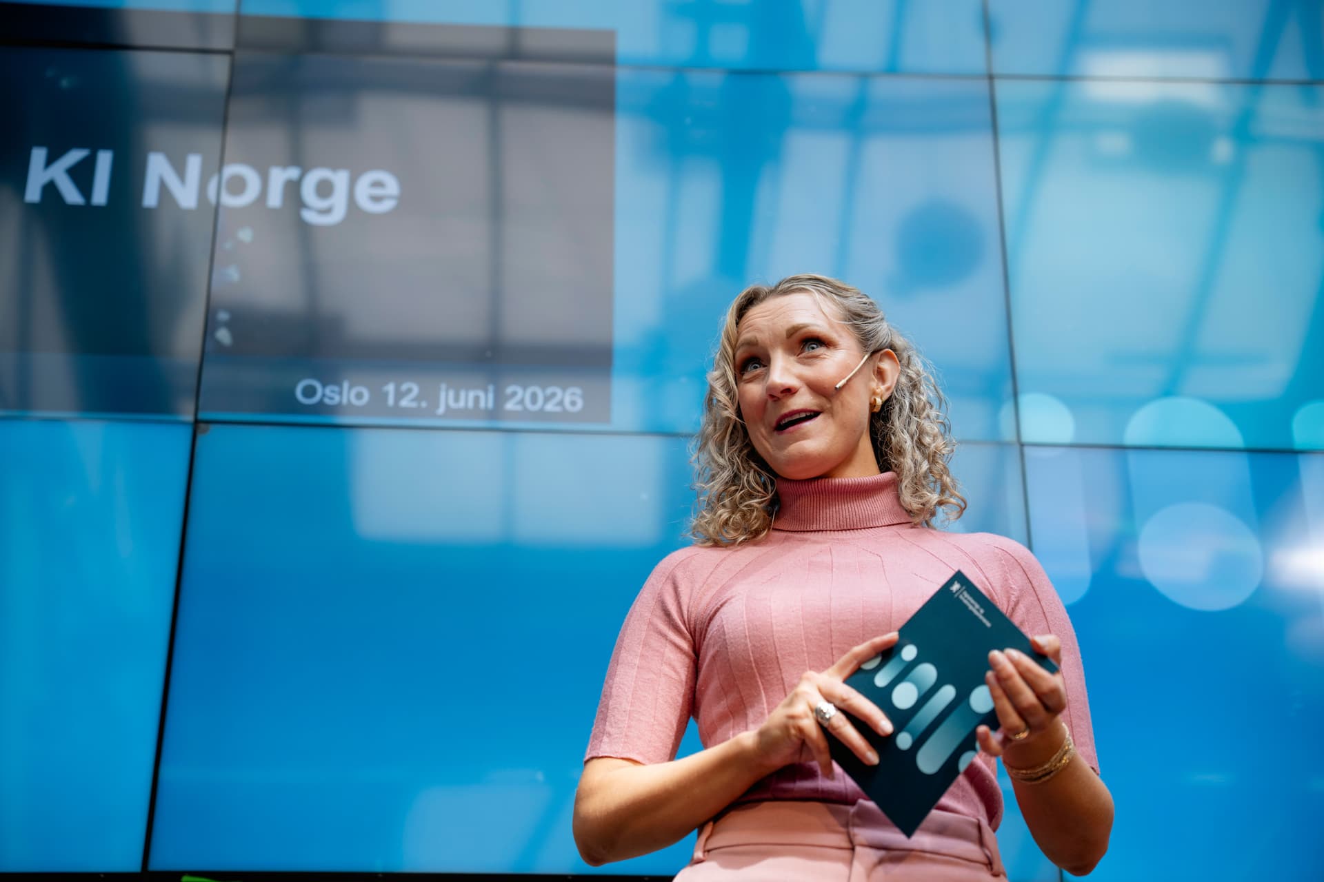 En kvinne med mikrofon snakker foran en blå skjerm med teksten «KI Norge» og «Oslo 12. juni 2026.»