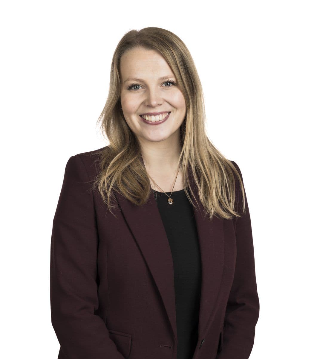 En kvinne med langt blondt hår, iført rødbrun blazer og svart topp, står smilende mot en hvit bakgrunn.