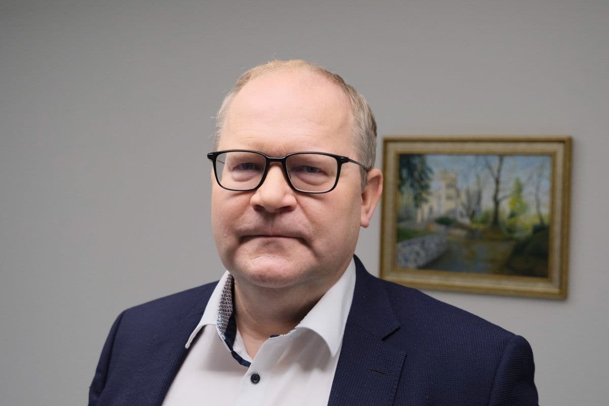 En middelaldrende mann med briller og nøytralt ansiktsuttrykk står innendørs foran en vegg med et innrammet landskapsmaleri.