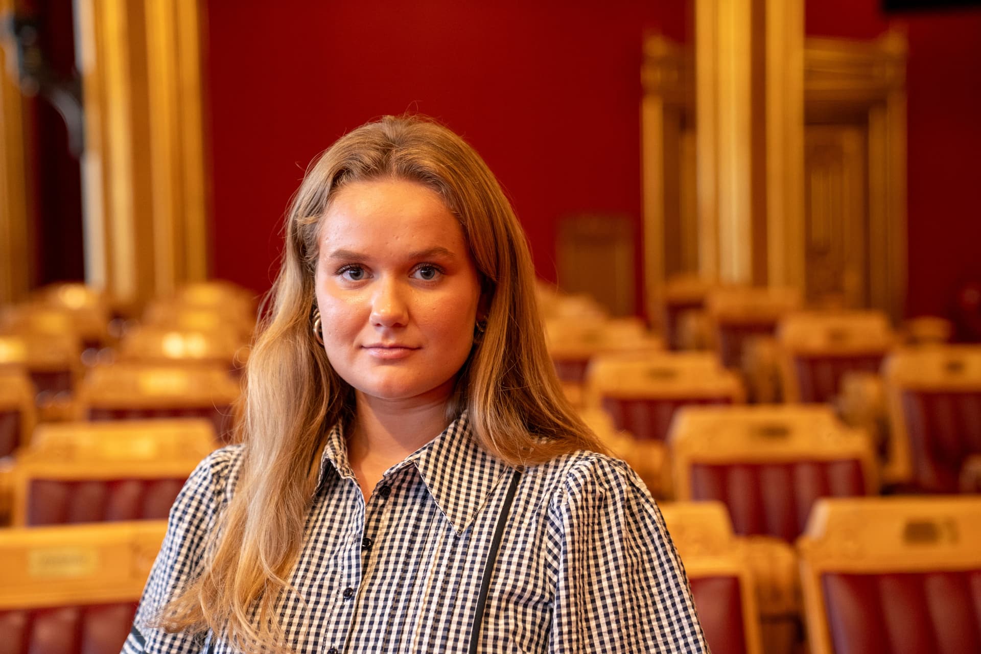En ung kvinne med langt blondt hår og rutete skjorte står i en tom, formell forsamlingssal.