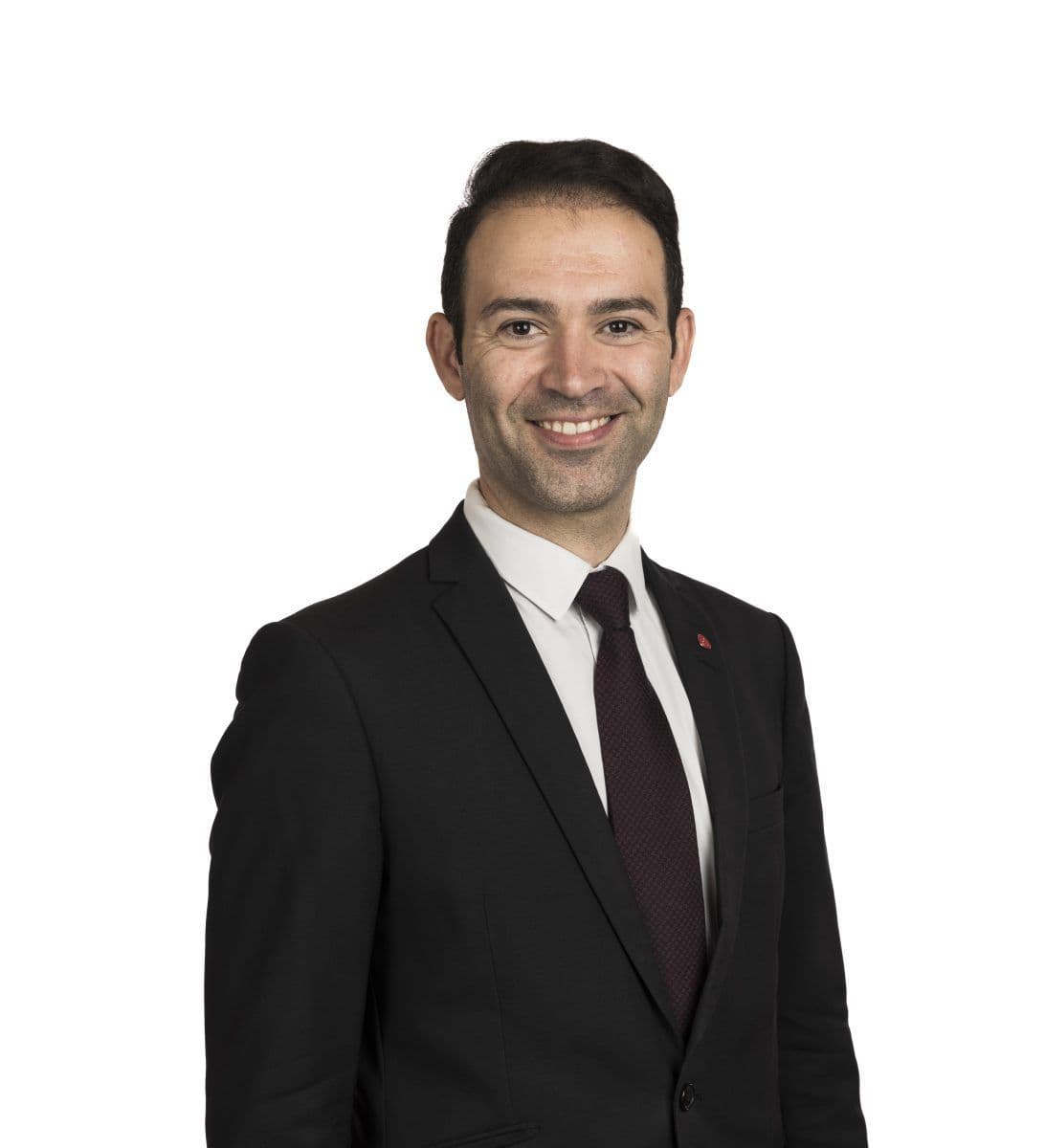 En mann i svart dress, hvit skjorte og mørkt slips smiler til kameraet mot en hvit bakgrunn.