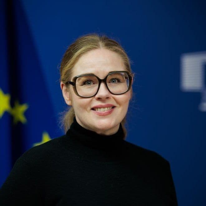 En kvinne med briller og svart rullekrage står foran et EU-flagg og et blått bakteppe med symboler fra Europakommisjonen.