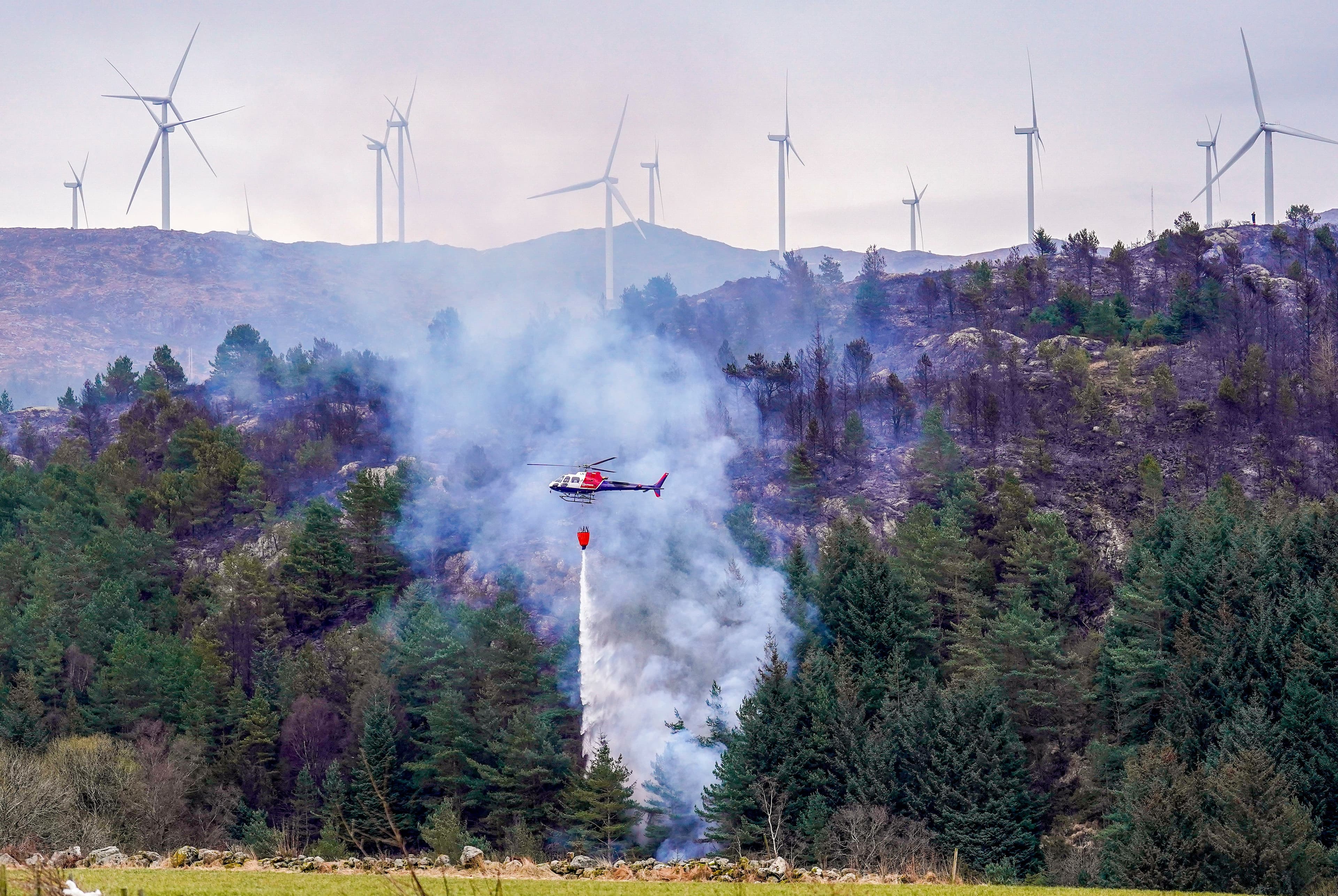 Et helikopter slipper vann over en røykfylt skogbrann, med vindturbiner på en ås i det fjerne.