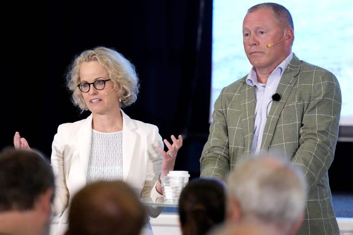 To personer, en kvinne og en mann, snakker på scenen foran et publikum. Begge har mikrofoner og forretningsantrekk; kvinnen gestikulerer mens hun snakker.