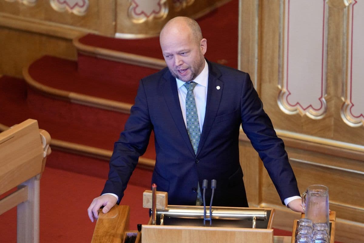 En mann i dress står på et podium i en formell sal og snakker inn i mikrofoner.