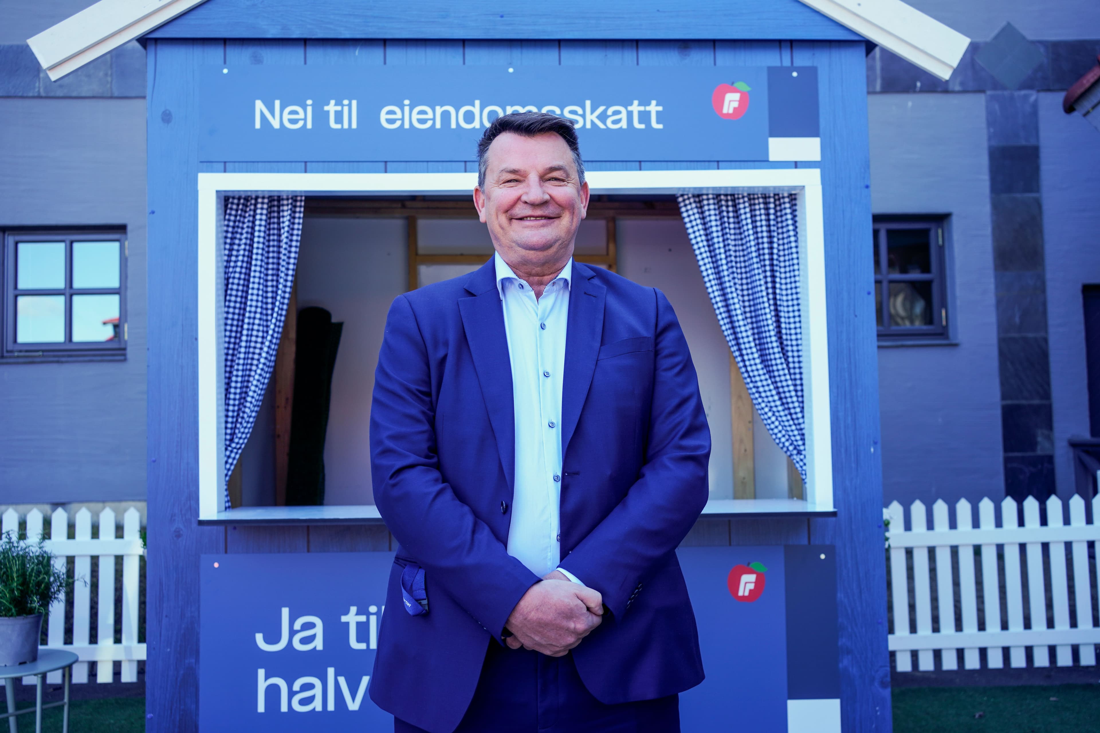 En smilende mann i blå dress står foran en blå politisk stand med teksten «Nei til eiendomsskatt» og en rød eplelogo.