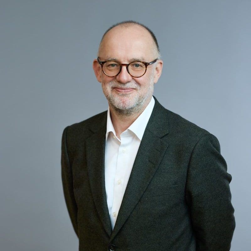 En mann med briller og skjegg, iført mørk dress og hvit skjorte, står mot en vanlig grå bakgrunn.