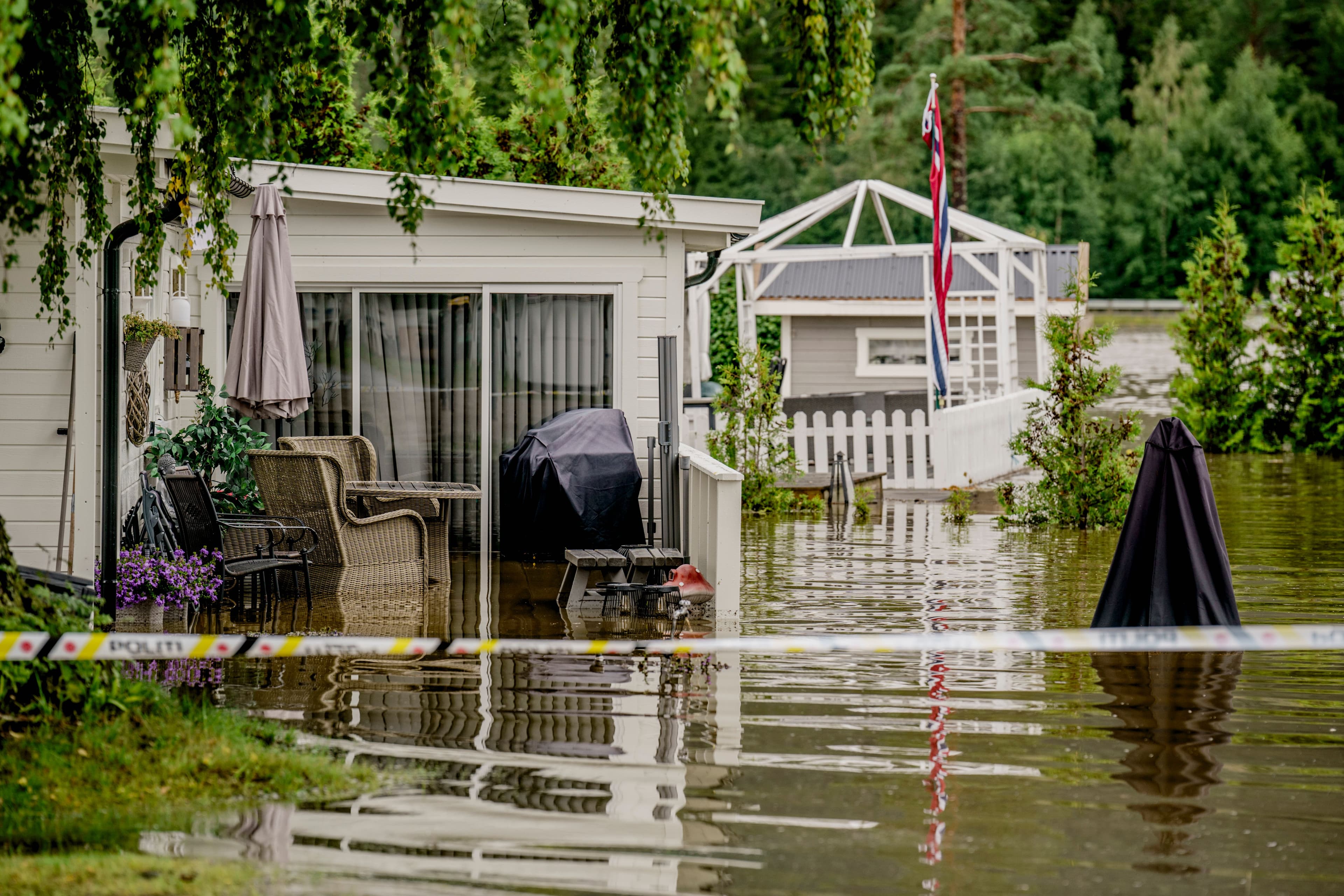 Flomvann omringer et hus og en terrasse og oversvømmer møbler, med sperrebånd merket «POLITI» synlig i forgrunnen.