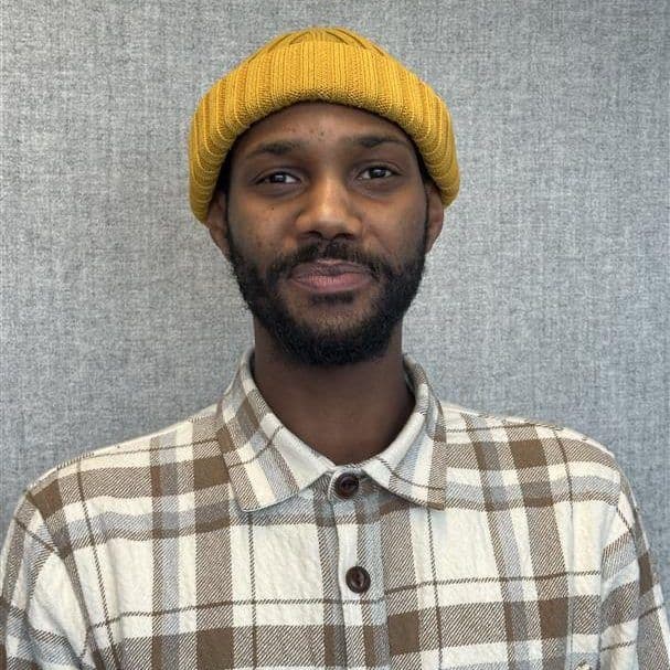 Person som har på seg en gul lue og rutet skjorte står mot en grå bakgrunn og smiler lett.