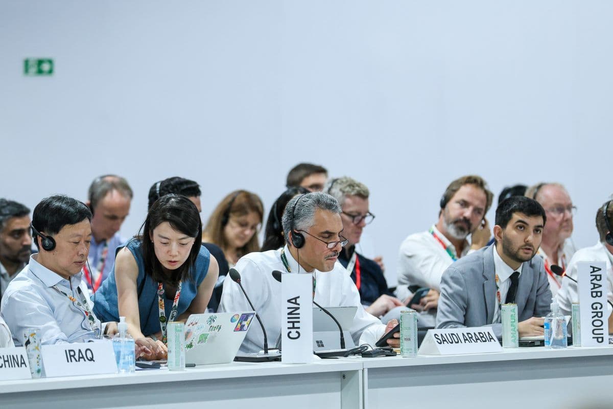 Delegater fra Kina, Irak, Saudi-Arabia og Den arabiske gruppen sitter ved et konferansebord med bærbare datamaskiner og dokumenter, iført hodetelefoner og lytter oppmerksomt.