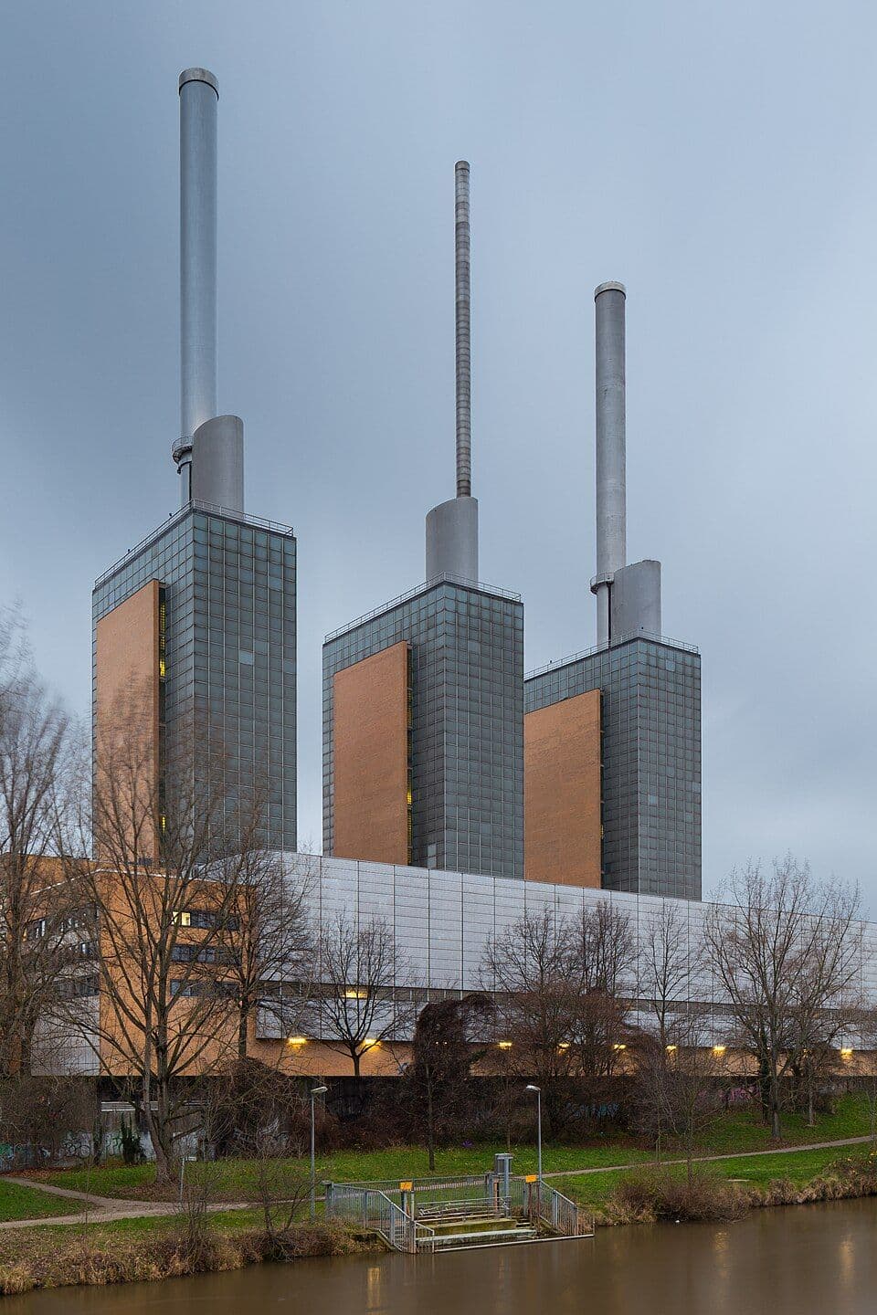 Three modern industrial buildings with tall smokestacks, featuring glass and reddish-brown facades, stand by a river under an overcast sky.