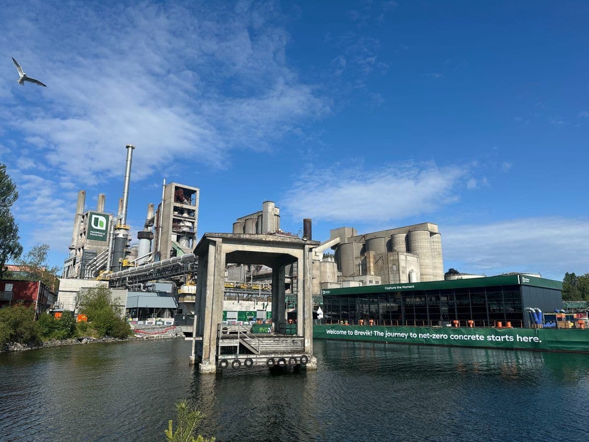 En stor sementfabrikk med siloer og industristrukturer i nærheten av et vann; på et grønt skilt står det: "The journey to net-zero concrete starts here." En måke flyr over bildet.