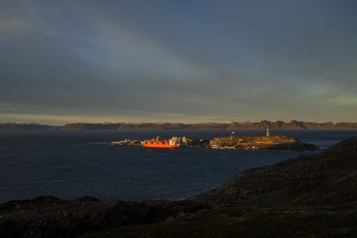 Et stort rødt skip ligger til kai ved et industrianlegg på en kystøy, med fjell og vann i bakgrunnen under en stort sett skyet himmel.